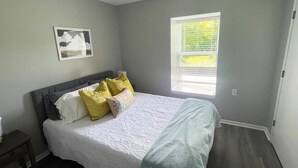 A bedroom with a bed, pillows, a nightstand, a window, and a picture on the wall. - The Ingrid - Entire 2 Bedroom Apt in Quilt Town, USA (Hamilton)