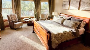 A bedroom with a large bed, two armchairs, a small table, and a view of a forest through large windows. - Riverfront Mansion with Indoor Pool, Private Pond, and Hot Tub on 16 Acre Ranch (Hamilton)