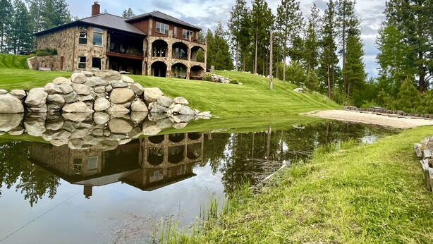 A large stone house with multiple windows and a stone wall reflecting in a pond. - Riverfront Mansion with Indoor Pool, Private Pond, and Hot Tub on 16 Acre Ranch (Hamilton)