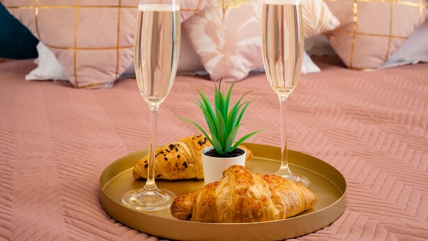 A bed with a pink quilt, a tray with croissants and champagne glasses, and a small plant.