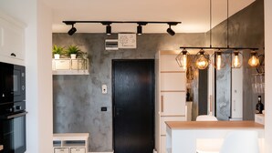 A modern kitchen with a black door, white cabinets, and a built-in oven. There are potted plants on a shelf, and pendant lights above the counter.