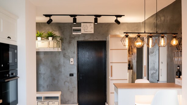 A modern kitchen with a black door, white cabinets, and a built-in oven. There are potted plants on a shelf, and pendant lights above the counter.