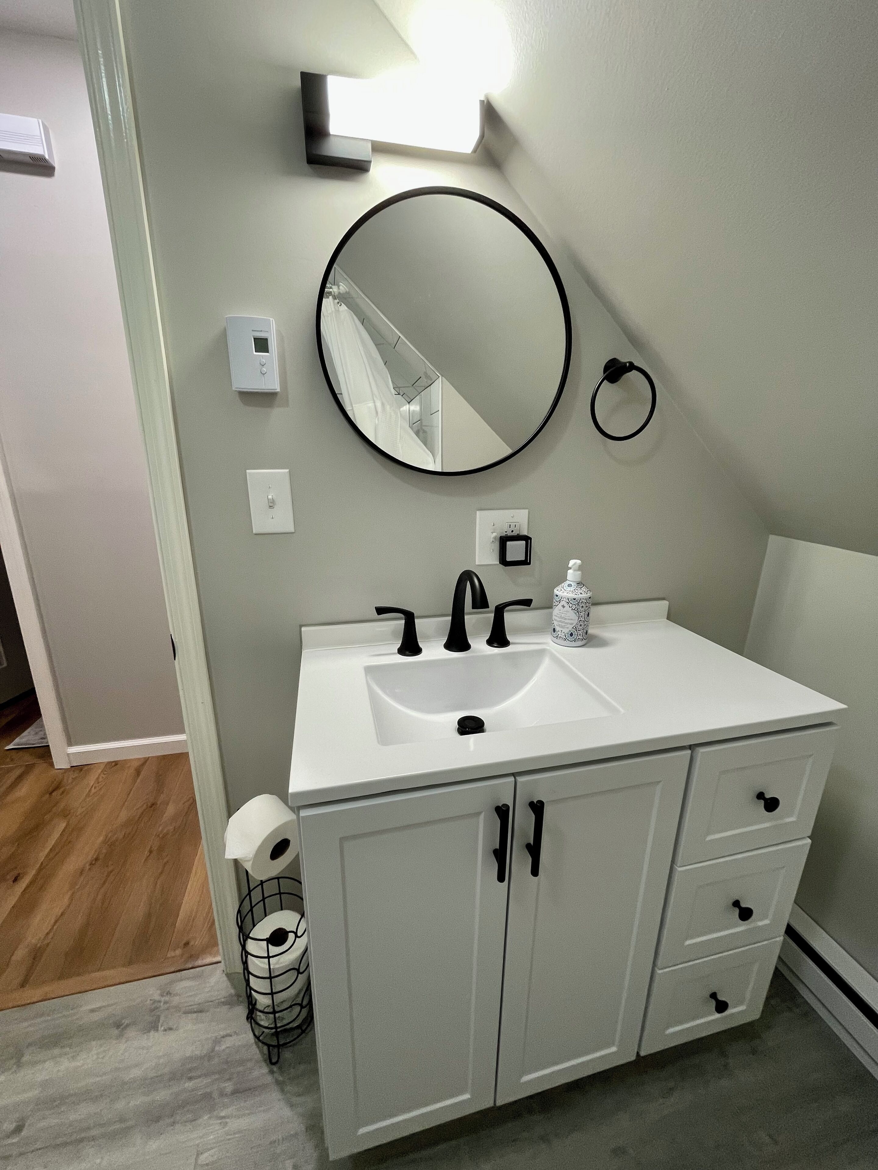 Een badkamer met een witte wastafel, een zwarte kraan en een ronde spiegel. Er is een toiletrolhouder en een lichtschakelaar op de muur.