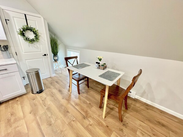 Un comedor con una mesa blanca, dos sillas de madera, una planta en maceta y un cubo de basura.