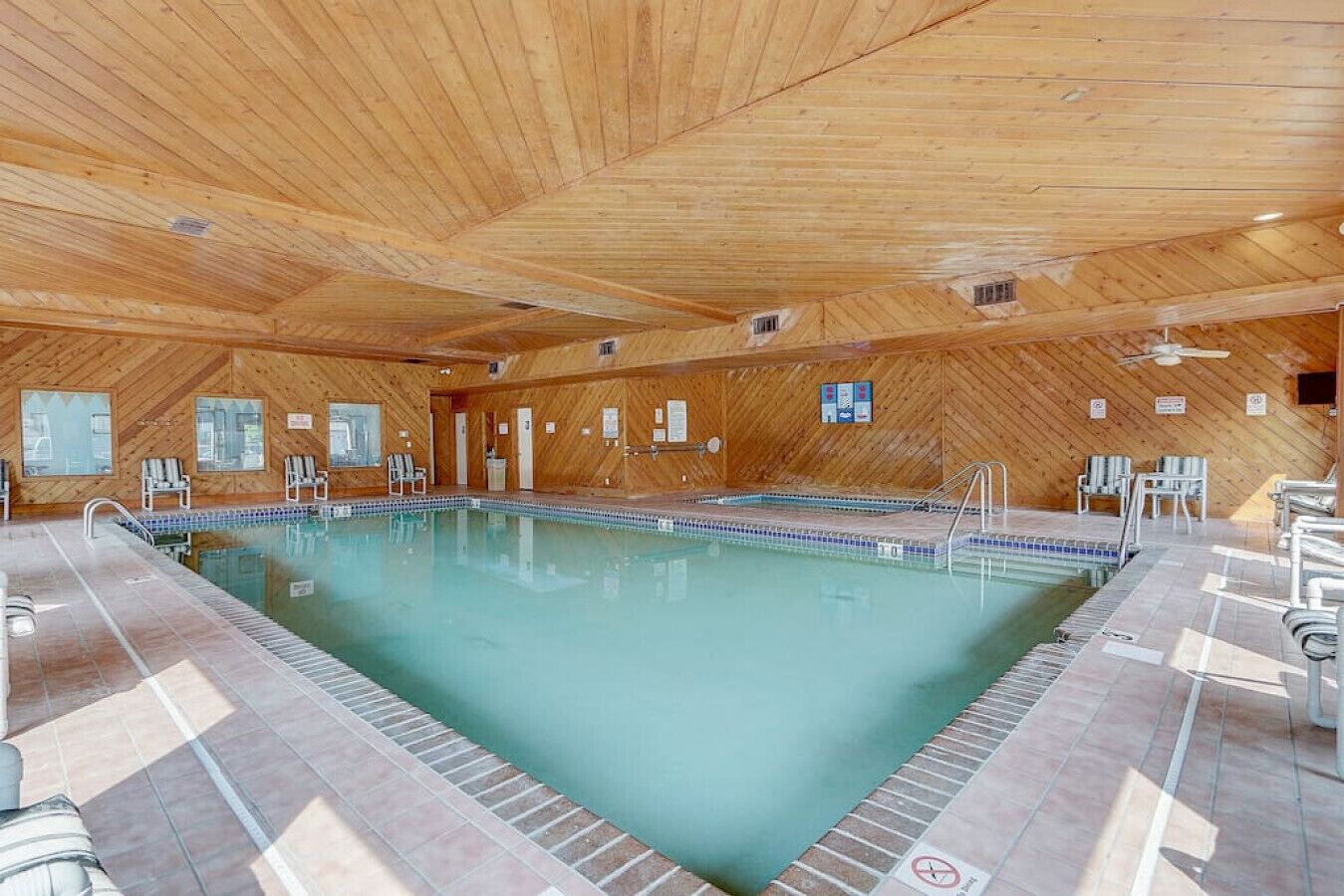 Een binnenzwembad met houten wanden en plafond, betegelde vloer en meerdere lounge fauteuils rondom het zwembad.