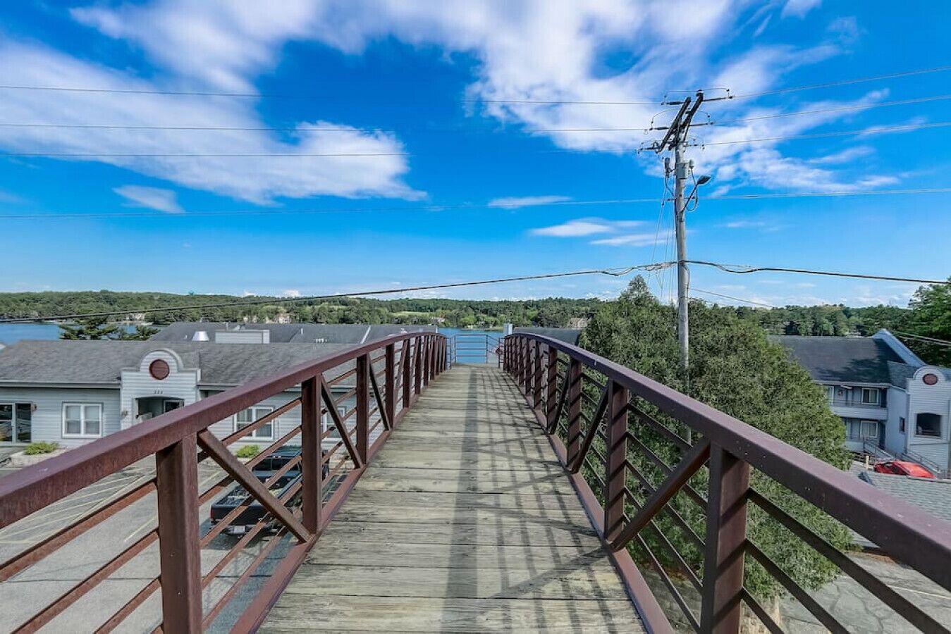 A pedestrian bridge with a metal railing, leading to a residential area with houses and a body of water in the background.