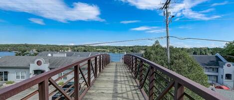 A pedestrian bridge with a metal railing, leading to a residential area with houses and a body of water in the background.
