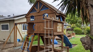 Een houten speelhuisje met een glijbaan, ladders en een raam.