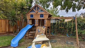 Een houten speelhuisje met een glijbaan en klimmuur, omgeven door een tuin.