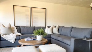 A modern living room with a grey sectional sofa, a round coffee table, and two white ottomans. There is a potted plant on the coffee table and a framed artwork above the sofa. - Newly Renovated from Top to Bottom! (Grand Lake)