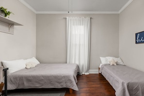 A bedroom with two beds, a window with sheer curtains, a wall-mounted shelf with a plant, and a blue sign with the word 'home'.