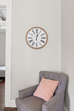 A room with a grey upholstered armchair, a wooden-framed wall clock, and a white wall.