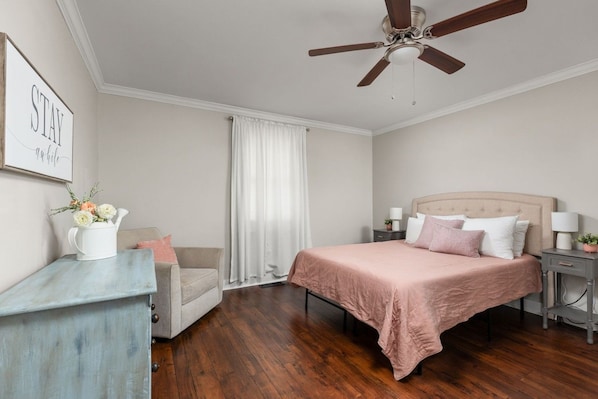 Une chambre à coucher comprenant un lit, une chaise, une table de chevet, un ventilateur de plafond et un mur orné d’un tableau.