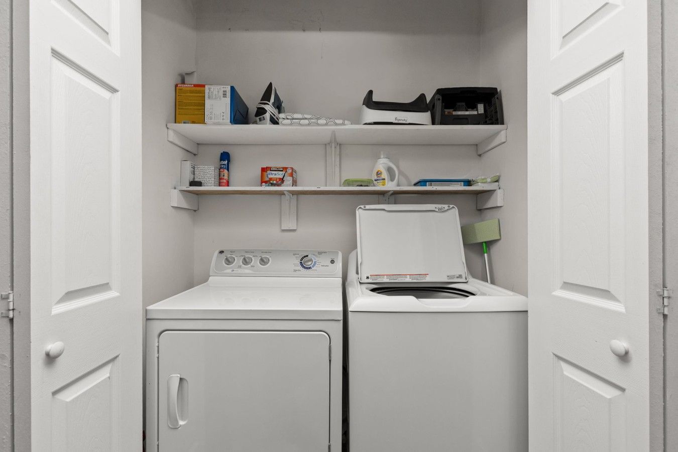A laundry room with a front-loading washer and dryer, shelves with cleaning supplies, and a broom.