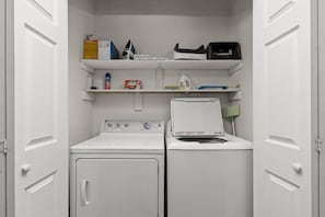 Une buanderie équipée d’un lave-linge et d’un sèche-linge à chargement frontal, d’étagères contenant des produits de nettoyage et d’un balai.