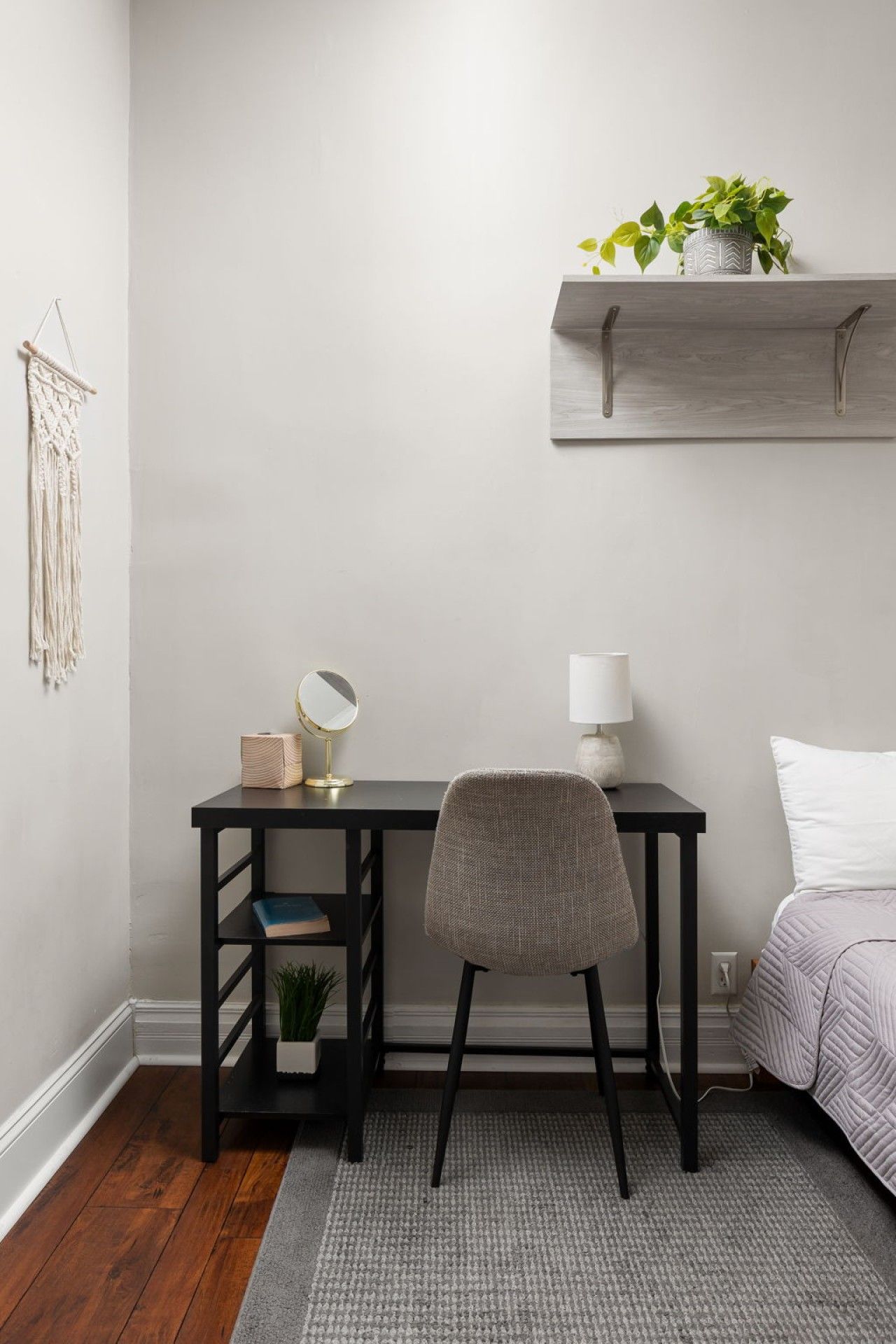 Une chambre avec un lit, un bureau avec une chaise, une table de chevet, une étagère murale agrémentée d’une plante et une décoration murale en macramé.