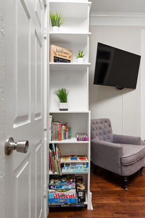 Une pièce avec une bibliothèque blanche, un fauteuil gris et un téléviseur fixé au mur.