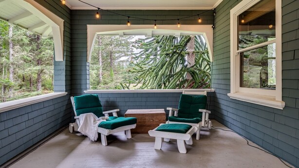 A covered porch with two white chairs and a table, a large window with a view of trees, and string lights on the ceiling. - Private Beach Estate 6B, Group Friendly (Pacific County)