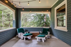 A covered porch with two white chairs and a table, a large window with a view of trees, and string lights on the ceiling. - Private Beach Estate 6B, Group Friendly (Pacific County)