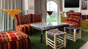 A living room with a sofa set, a coffee table, and a wooden chair. There is a large window with curtains, a flat-screen TV mounted on the wall, and a painting above the TV. - Lovely and clean Apartment (TANGER)
