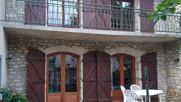 A two-story stone building with wooden shutters, a balcony with metal railings, and a stone patio with outdoor furniture.