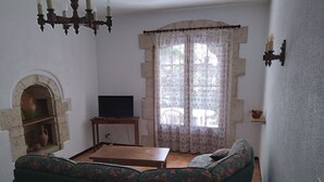A living room with a sofa, a television on a wooden stand, a stone wall, and a window with lace curtains.