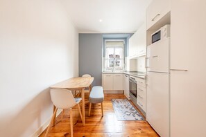 Een smalle keuken met een houten tafel en stoelen, een raam met gordijnen en een koelkast.