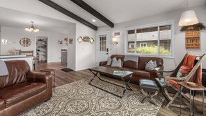 A spacious living room with a brown leather sofa, a patterned rug, a coffee table, and a rocking chair. There is a kitchen in the background with a white door, and a window with blinds. - Immaculate 3 bedroom with hot tub , AC , close to golf course , Whitefish lake , Whitefish Trails! (Whitefish)