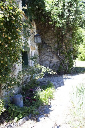 A stone building with a narrow pathway leading to a door, surrounded by lush greenery and flowering plants.