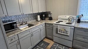 Una cocina moderna con armarios grises, microondas, cafetera y hervidor. Hay dos alfombras en el suelo con frases relacionadas con comida.