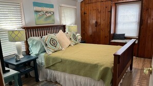 Un dormitorio con cama, mesitas de noche, un armario de madera, una ventana con persianas y un cuadro en la pared.