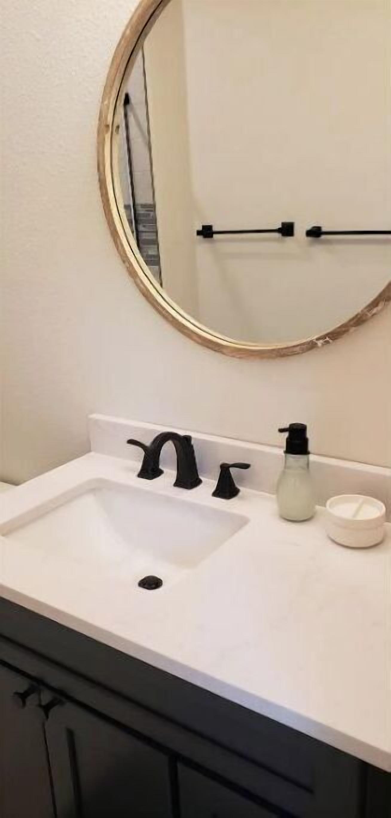Une salle de bain avec un lavabo blanc, un robinet noir et un miroir rond doté d’un cadre doré.