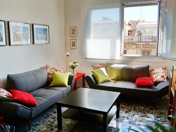 Un salón con un sofá gris, una mesa roja y una mesa de centro negra. Hay dos ventanas que dan a edificios.