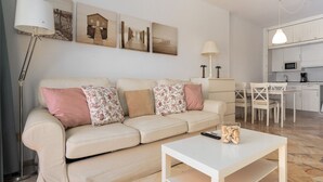 A living room with a beige sofa, a white coffee table, and a kitchen area with white cabinets and a dining table.