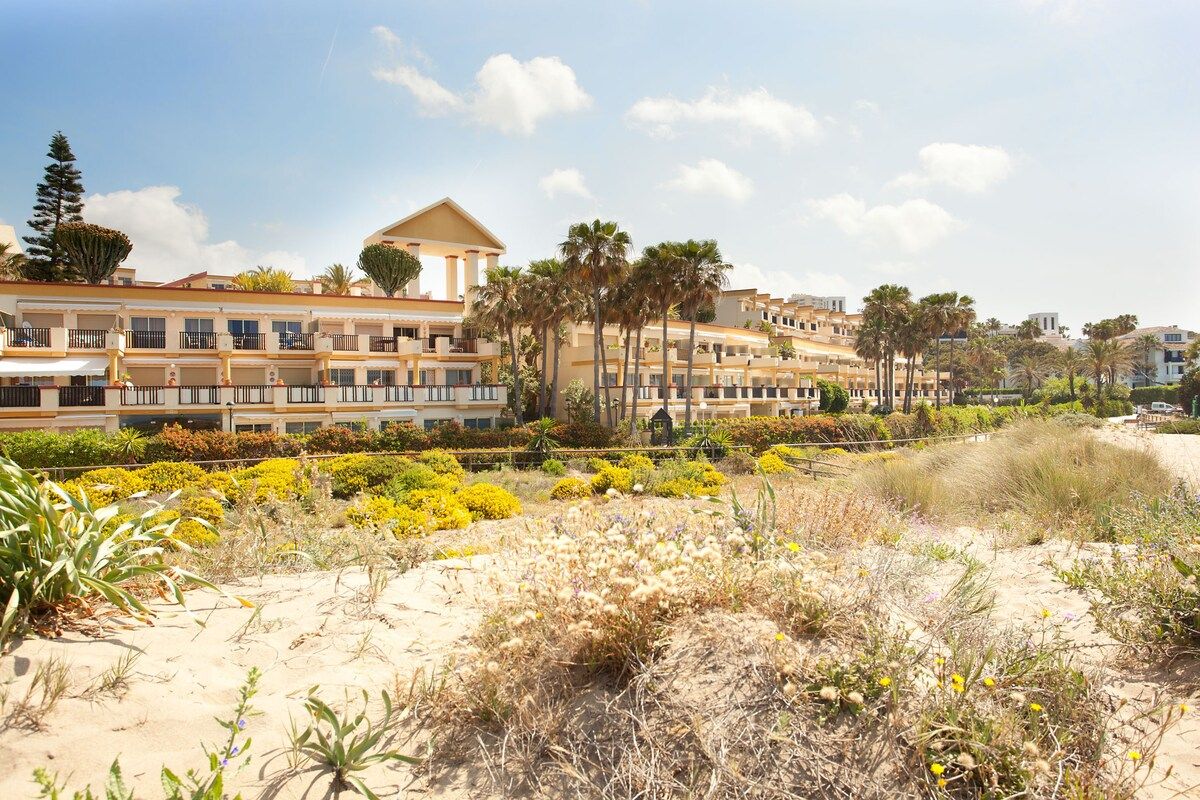 Un hotel frente al mar con varios balcones, una zona de playa arenosa y diversa vegetación.