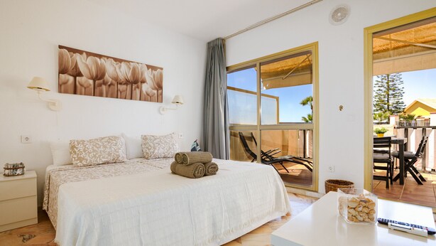 A bedroom with a bed, a nightstand, a table with a glass container, and a large window with a view of the outside.