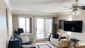 A living room with a sofa, a coffee table, a TV, and a balcony with a view. - Oceanfront 3-BR boardwalk condo w indoor pool! (Virginia Beach)