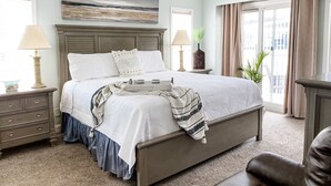 A neatly arranged bedroom with a large bed, bedside lamps, a nightstand, a dresser, and a view of the outdoors through a window. - Oceanfront 3-BR boardwalk condo w indoor pool! (Virginia Beach)