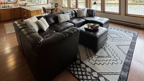 A spacious living area with a large black leather sectional sofa, a coffee table, and a patterned rug. There are multiple windows providing natural light and views of the outdoors. - Riverfront Cabin! BBQ, HOT TUB, FIRE PIT, 4 BEDROOMS (Baring)