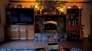 A cozy living room with a stone fireplace, a flat-screen TV, wooden cabinets, and a bookshelf. - Riverfront Cabin! BBQ, HOT TUB, FIRE PIT, 4 BEDROOMS (Baring)