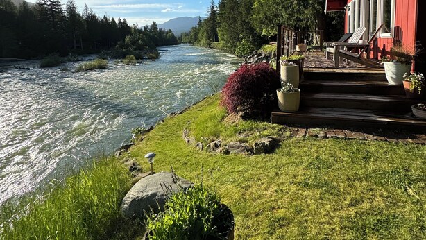 A riverside view with a red building, a wooden deck, and a river. - Riverfront Cabin! BBQ, HOT TUB, FIRE PIT, 4 BEDROOMS (Baring)