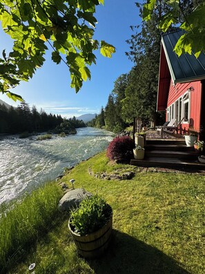 A riverside view with a red building, a wooden deck, and a river. - Riverfront Cabin! BBQ, HOT TUB, FIRE PIT, 4 BEDROOMS (Baring)