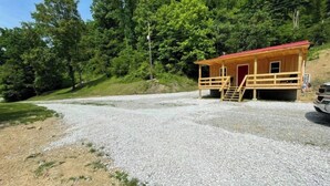 A small wooden cabin with a red roof, a gravel driveway, and a forested hillside in the background. - Country Charm (Williamson)