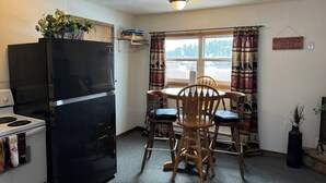 A compact kitchen with a black refrigerator, wooden dining table, and chairs. A window with curtains is visible, and a sign on the wall reads "What happens at the cabin stays at the cabin." - The fishing suite (Red Feather Lakes)