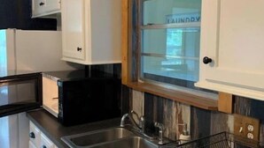 Une cuisine avec des meubles blancs, un évier en acier inoxydable, un four à micro-ondes et une fenêtre donnant sur une buanderie.