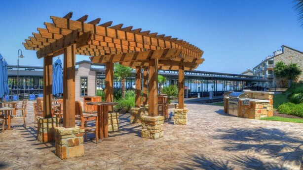 Een terras buiten met een houten pergola, stenen zitplaatsen en uitzicht op een gebouw aan het water.