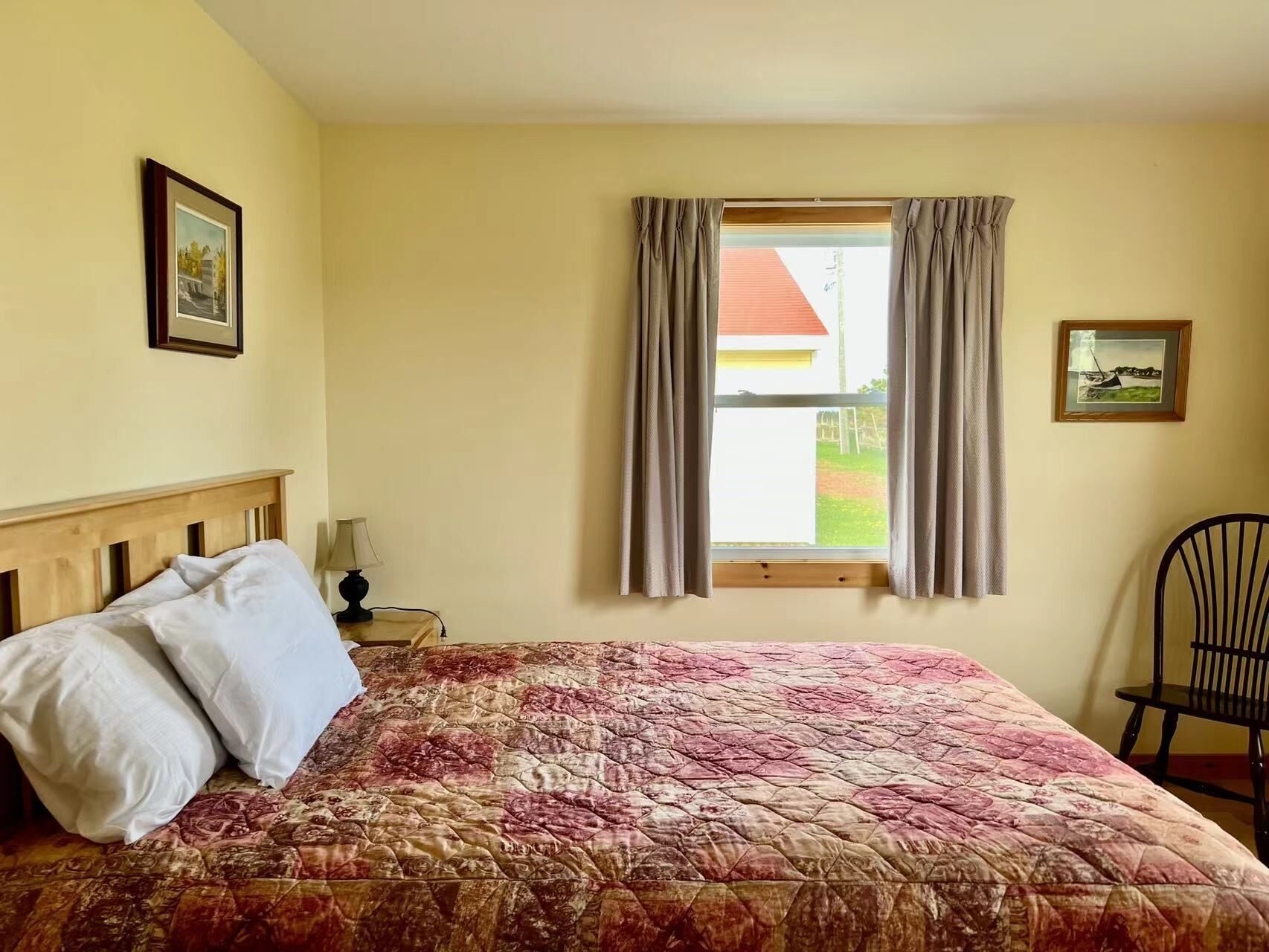 A bedroom with a bed, a window with curtains, a chair, and two framed pictures on the wall.