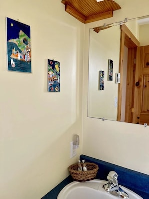 Un baño con un lavabo blanco, un espejo y dos obras de arte en la pared que representan escenas coloridas.