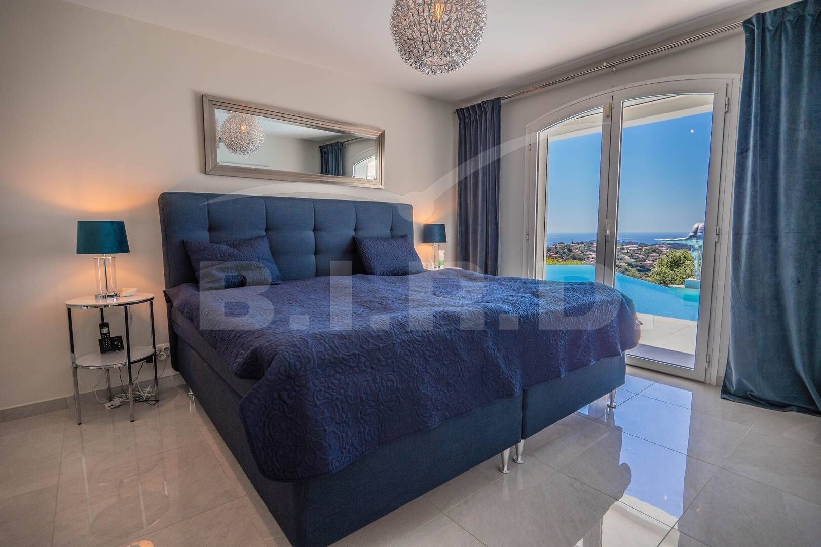 A modern bedroom with a large bed, a mirror above, and a view of the sea through a sliding glass door.