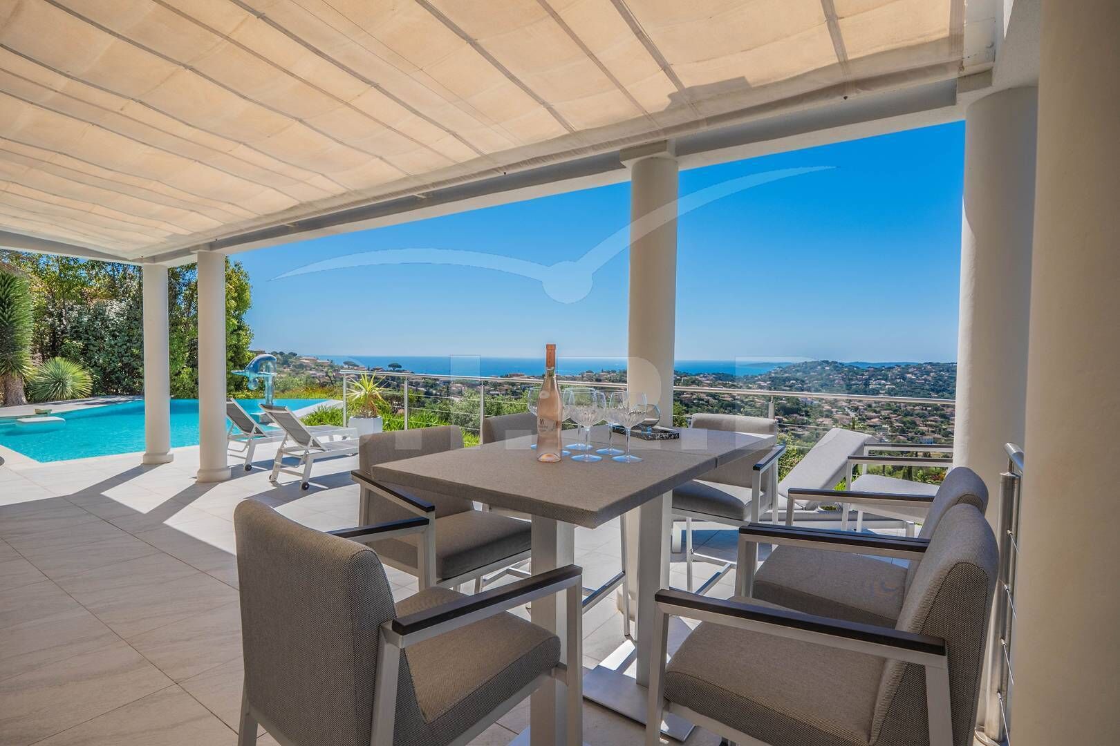 A covered outdoor seating area with a pool, chairs, and a table with wine glasses.